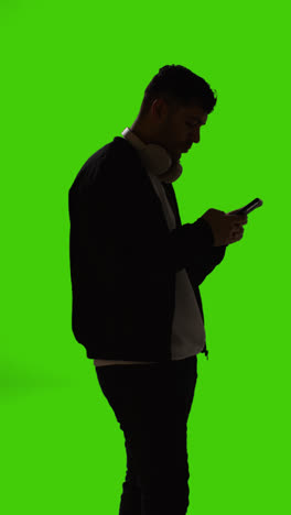 Vertikale-Videosilhouette-Eines-Mannes-Mit-Kabellosen-Kopfhörern,-Der-Textnachrichten-Auf-Dem-Mobiltelefon-Vor-Einem-Grünen-Bildschirm-Sendet